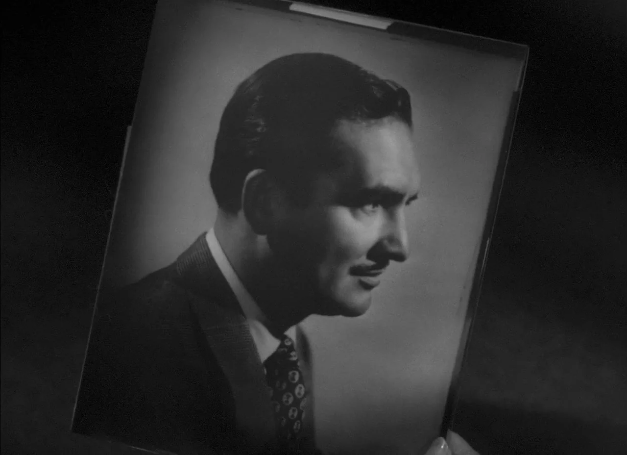 The Amazing Mr. X (1948) movie still: closeup — A close-up shot displays a handheld framed black-and-white portrait of a man. The man in the…