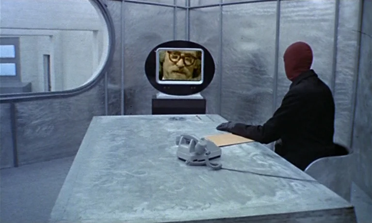 Nuits Rouges (1974) movie still: medium — A room with silver-colored, textured walls and a large, flat gray table. A person wearing a red mask sits…
