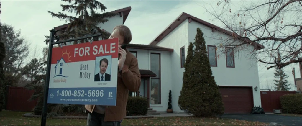 Clown (2014) movie still: medium — A man stands in front of a white two-story suburban home with brown garage doors. He holds a real estate sign…