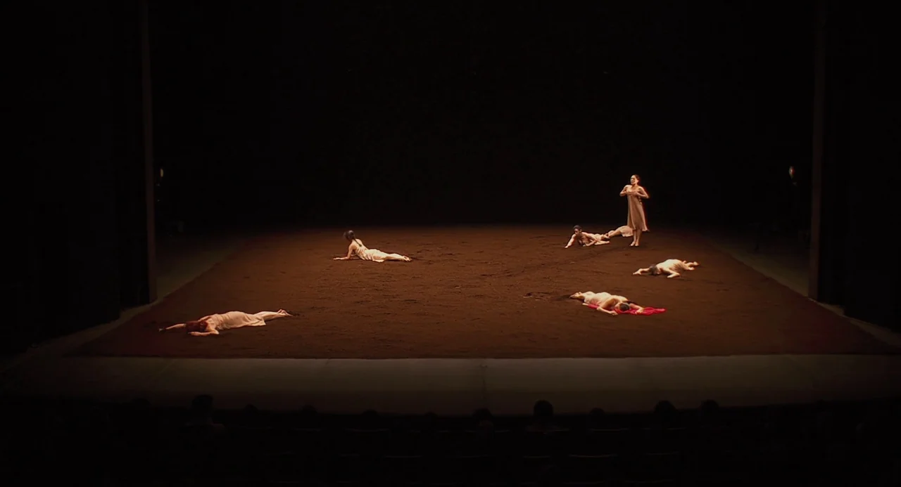 Pina (2011) movie still: wide — A wide shot shows a performance stage covered in a thick layer of dark soil. Several dancers in beige dresses lay or…