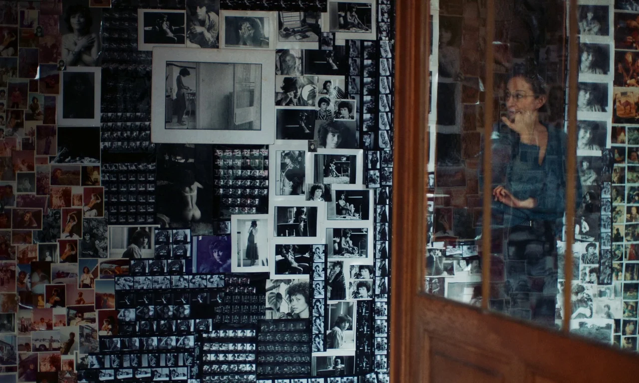 Little Girl Blue (2023) movie still: medium — A wall covered in numerous monochrome photographs and contact sheets stands beside a glass door. A…