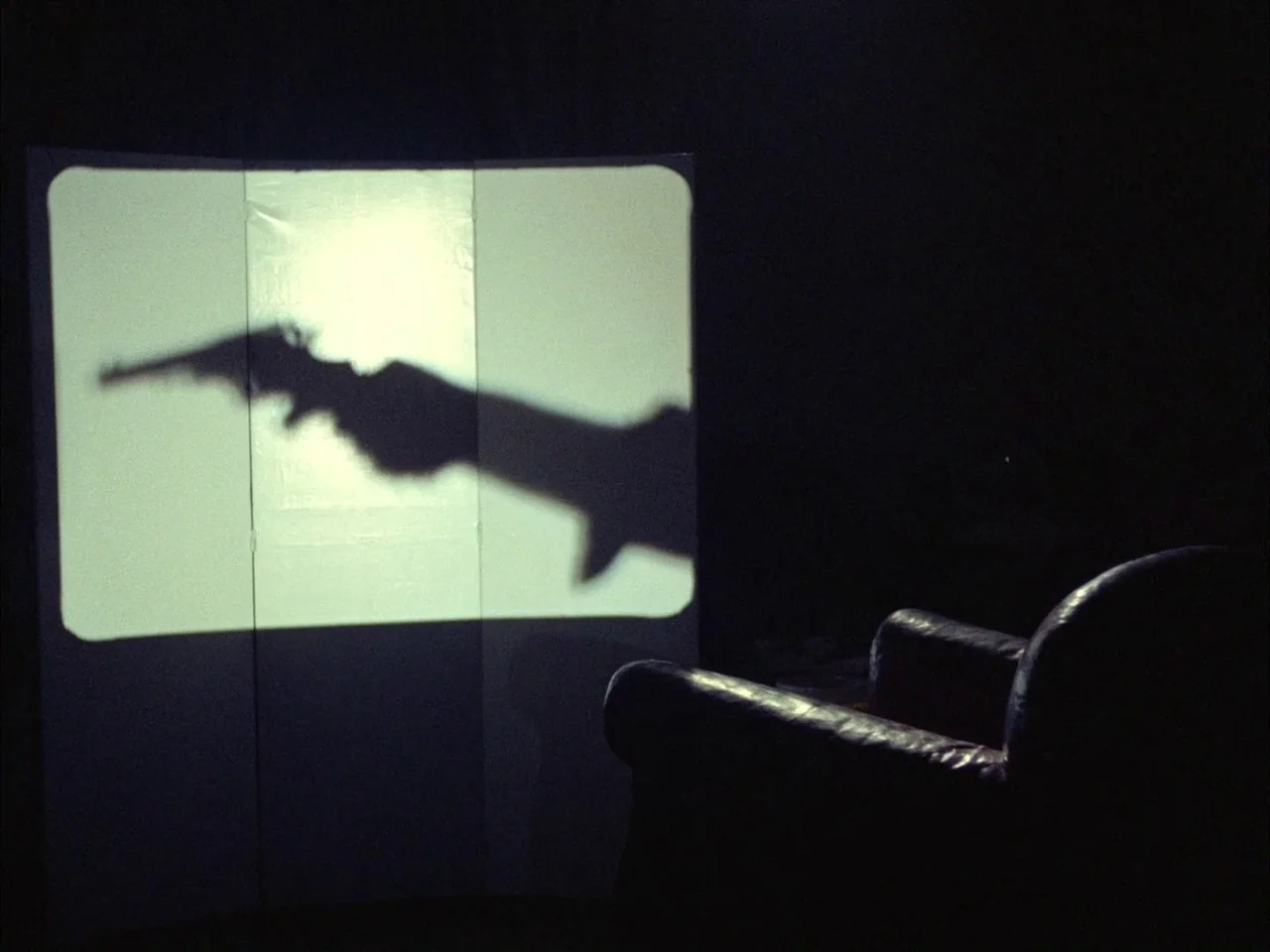 Dillinger is Dead (1969) movie still: wide — A dark, empty room contains a white partition screen and a dark leather armchair. A silhouette of a…