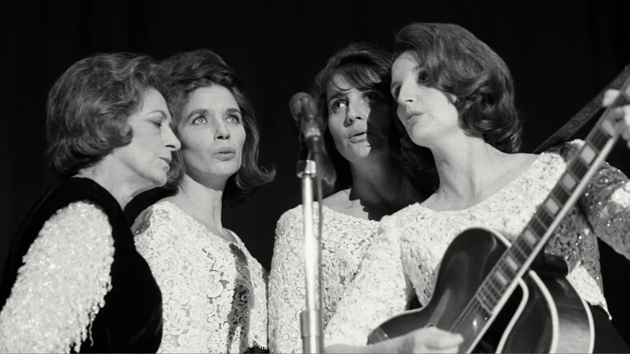 The Winding Stream (2014) movie still: medium — A dimly lit indoor music performance space with a black curtain backdrop. Four women stand in a row…