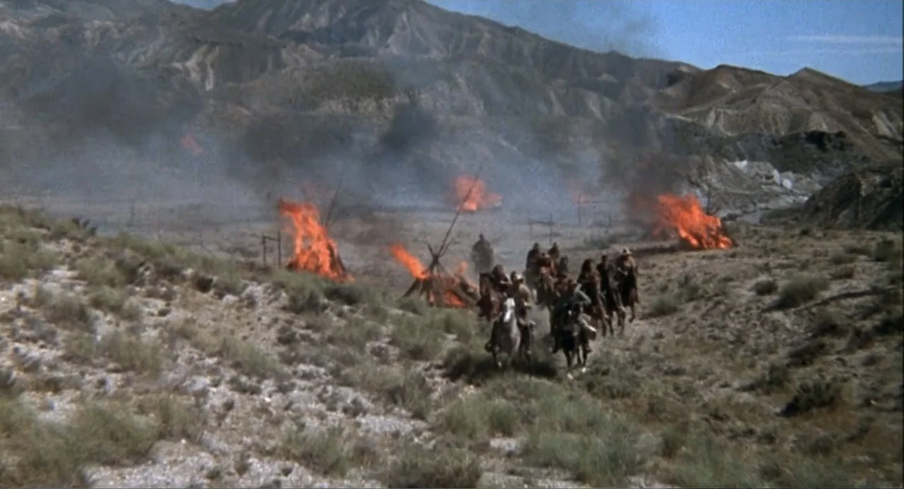 Chato's Land (1972) movie still: wide — A dry desert valley features a village of wooden structures engulfed in large orange flames. A small group…
