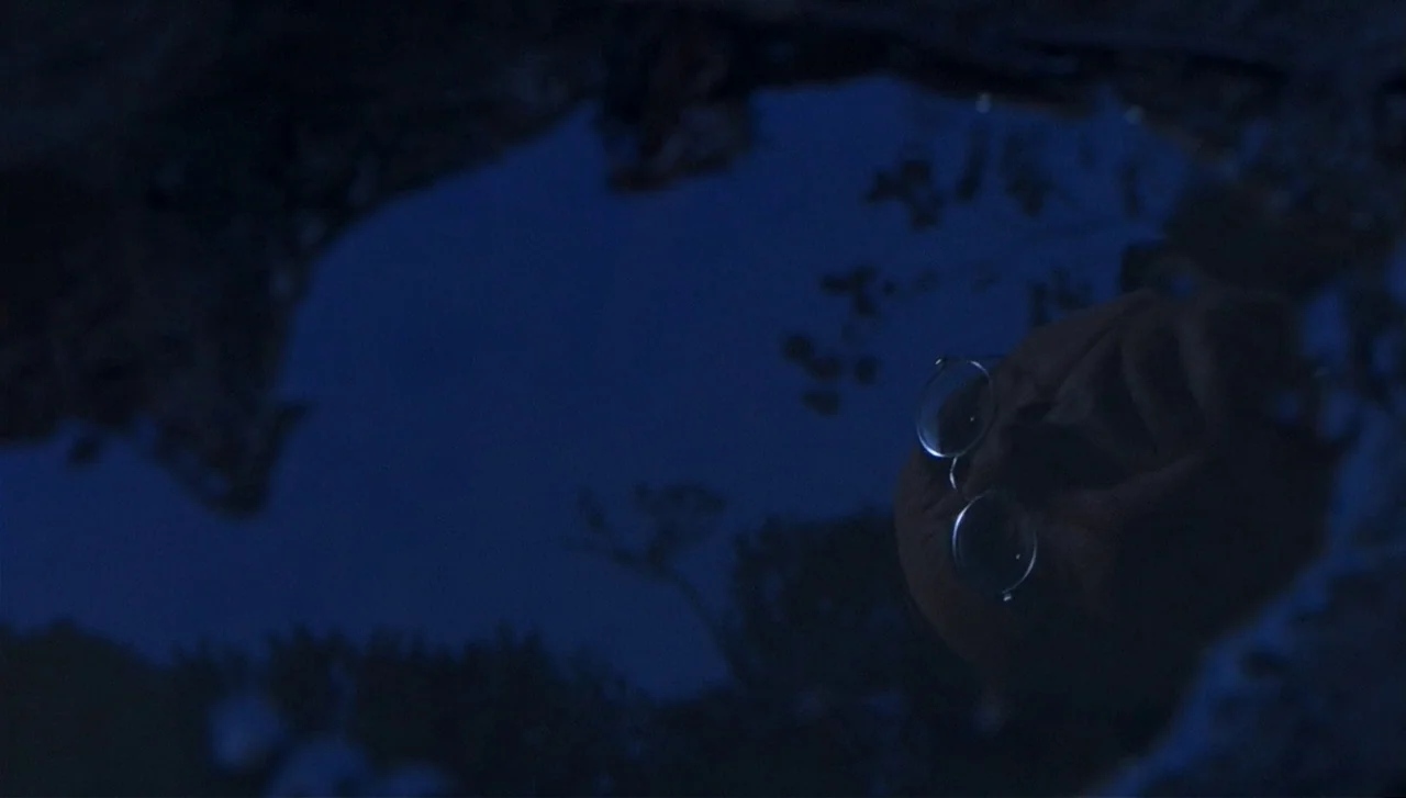 The Lost World: Jurassic Park (1997) movie still: closeup — A dark water surface reflects the face of a person wearing round glasses. The…