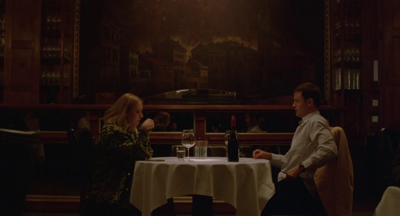 Sick of Myself (2022) movie still: wide — A dimly lit restaurant features dark wood paneling and a large, shadowed mural background. A man and a…