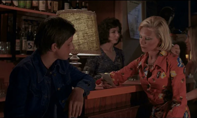 Small Change (1976) movie still: medium — A dimly lit tavern interior features a wooden counter and a central lampshade. A blonde woman in a floral…