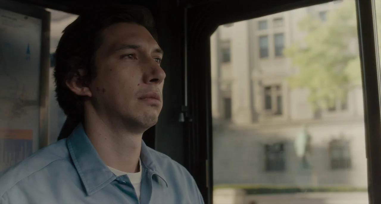 Paterson (2016) movie still: medium — A man sits inside a transit vehicle next to a large clear window. Outside the window, a stone building and…