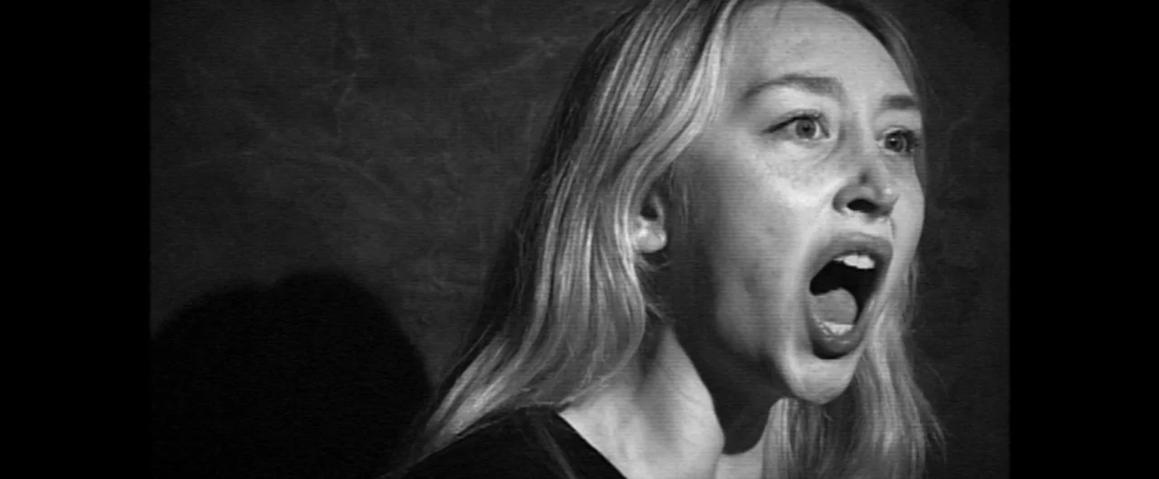 Poser (2021) movie still: closeup — An interior space features a textured, dark wall serving as a plain background. A young woman with blonde hair…