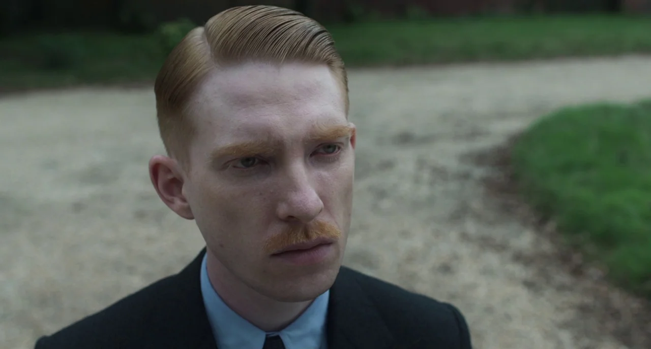 The Little Stranger (2018) movie still: closeup — An outdoor setting with a light-colored gravel path and patches of green grass nearby. A man with…