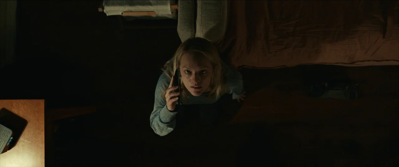 The Invisible Man (2020) (2020) movie still: medium — A dimly lit room with dark floors and a bed partially visible in the frame. A woman looks…
