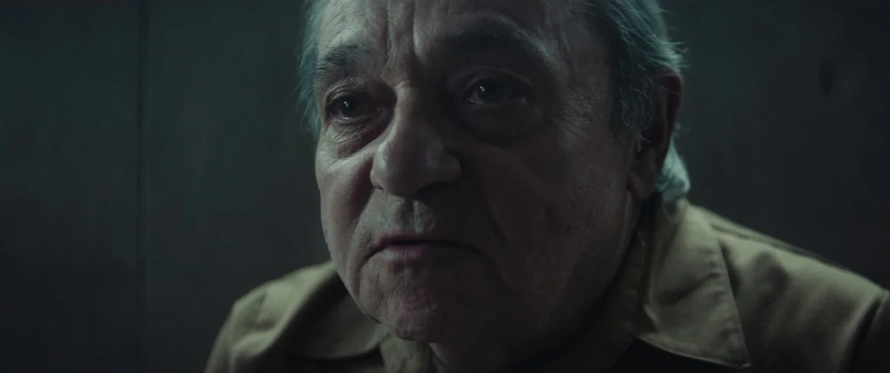 The Platform (2019) movie still: closeup — A dimly lit room with plain walls in neutral shades. An elderly man with graying hair sits and looks…