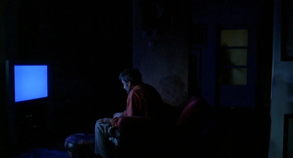 Uzak (2002) movie still: medium — A dark living room contains a man sitting alone on a couch. He faces a television set that casts a bright blue glow.