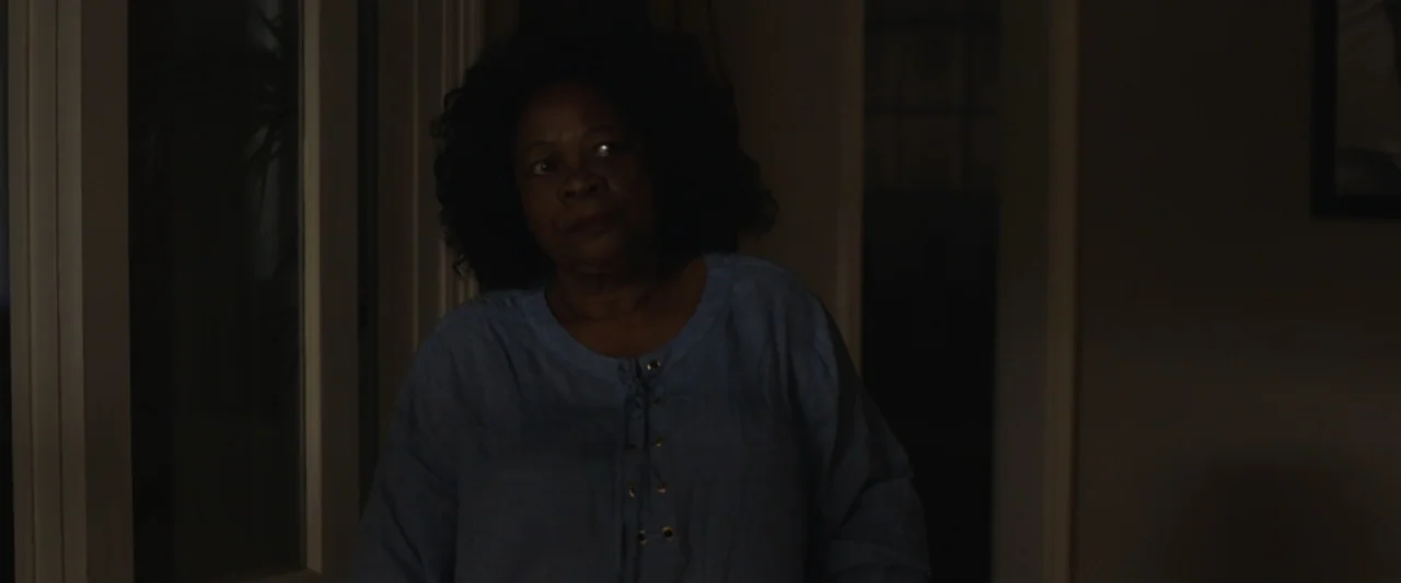 Mia and the White Lion (2018) movie still: medium — A dimly lit indoor hallway with dark walls and a glimpse of an adjacent room. An older woman in…