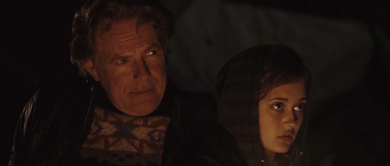 Wildlike (2014) movie still: medium — An outdoor nighttime scene illuminated by a nearby fire. An older man and a young woman sit together looking…
