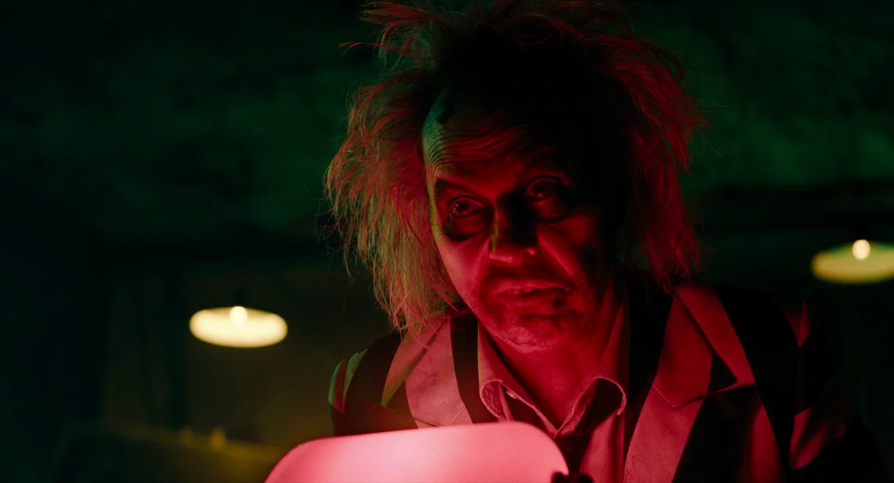 Beetlejuice Beetlejuice (2024) movie still: closeup — A dark, dimly lit room features low-hanging circular lights in the background. A man with wild…