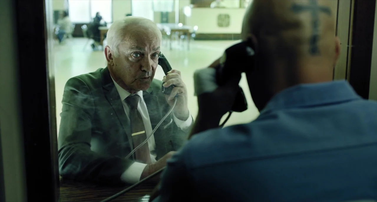 Brawl in Cell Block 99 (2017) movie still: medium — A man in a suit speaks through glass to a bald prisoner in uniform. They communicate via…