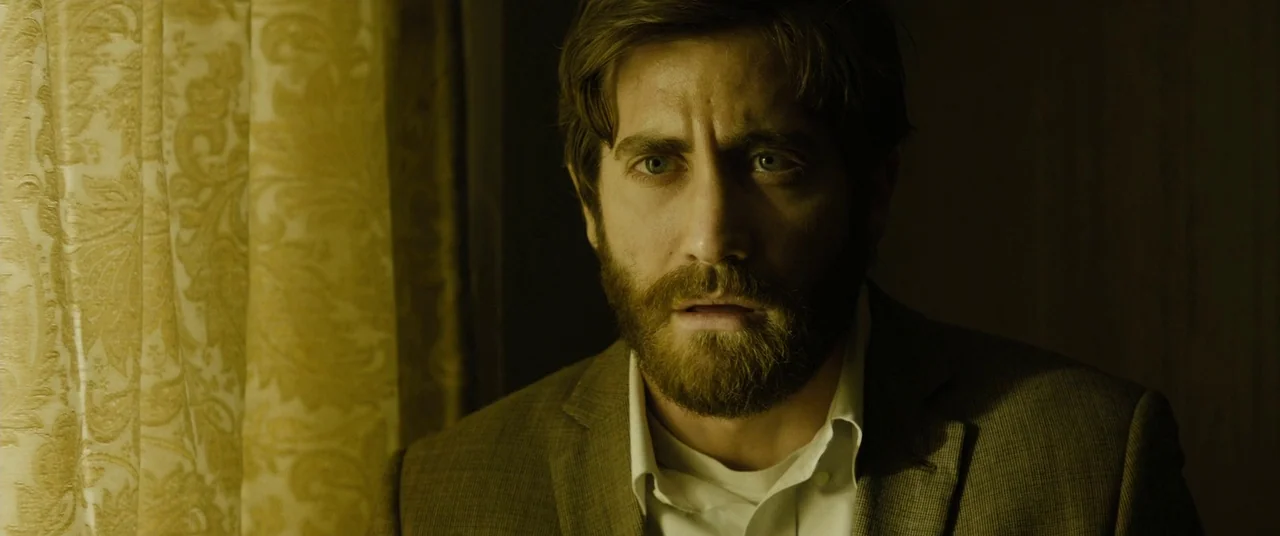 Enemy (2013) movie still: closeup — A dimly lit room contains a man standing near patterned golden curtains. He looks toward the side with an alert…