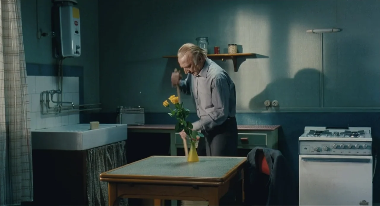 Le Havre (2011) movie still: medium — A utilitarian kitchen features a sink unit, a stove, and a small dining table. An elderly man sweeps the floor…