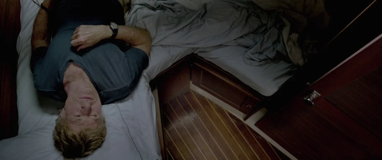 All Is Lost (2013) movie still: medium — A small yacht cabin features polished wood floor planks and a narrow white mattress. A man lies on his back…