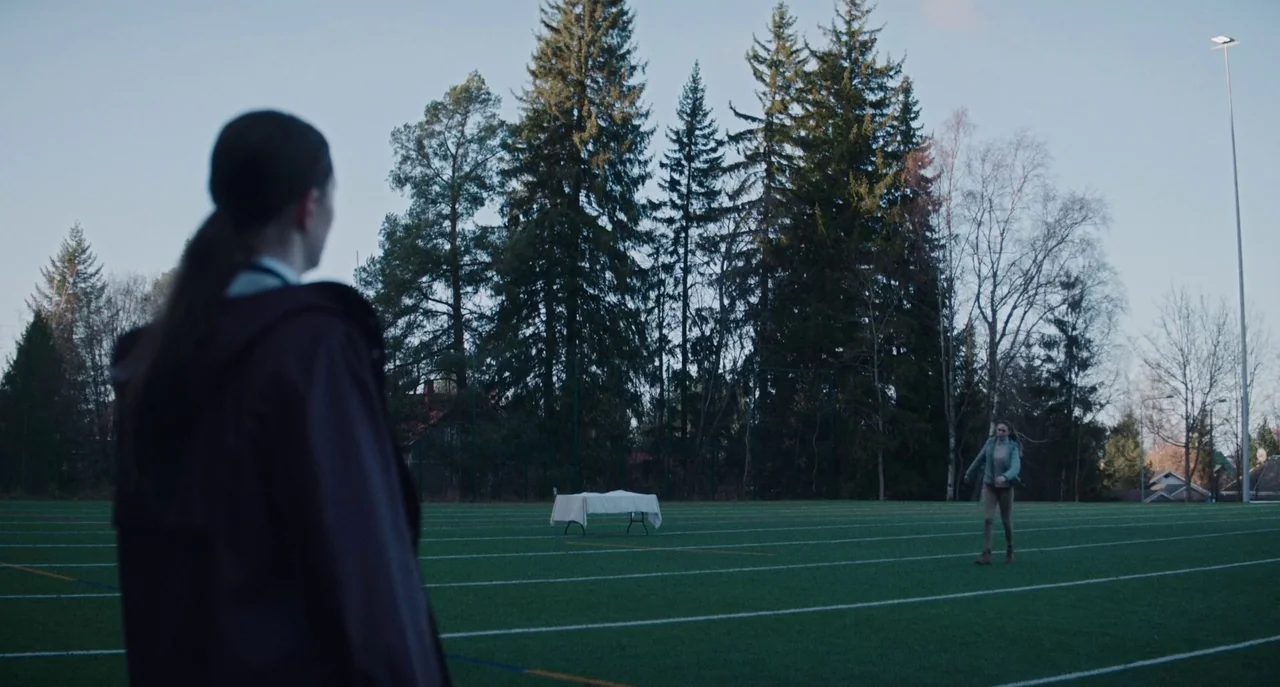 Dual (2022) movie still: wide — A synthetic turf field is surrounded by tall pine trees under a clear sky. A small folding table draped with a white…