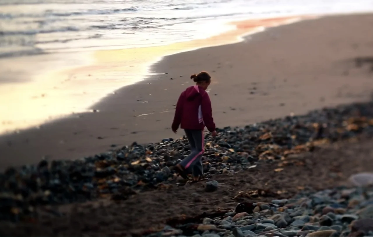 No Place to Hide (1992) movie still: fullbody — A rocky beach stretches along the water at golden hour with wet sand. A girl walks along the pebbles…