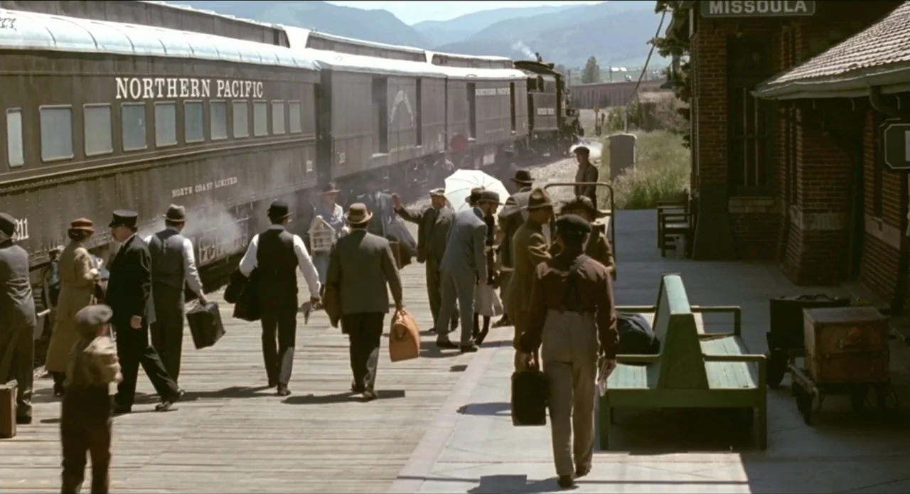A River Runs Through It (1992) movie still: wide — A bustling wooden train station platform features a large steam train idling at tracks. Various…