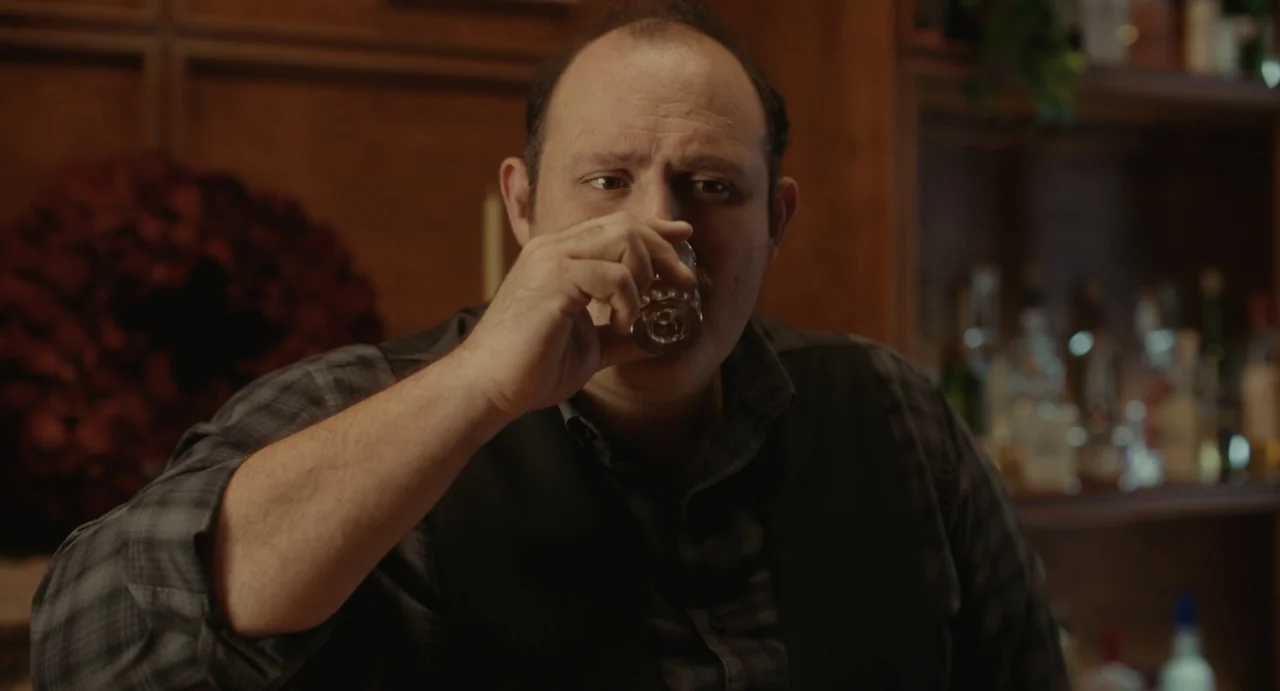 Dating Santa (2023) movie still: medium — A dimly lit bar setting with dark wood shelves holding glass bottles behind. A man with a balding head and…