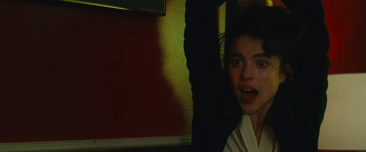 Sanctuary (2023) movie still: closeup — A dimly lit room features a deep red wall with a framed picture. A young woman with curly dark hair raises…