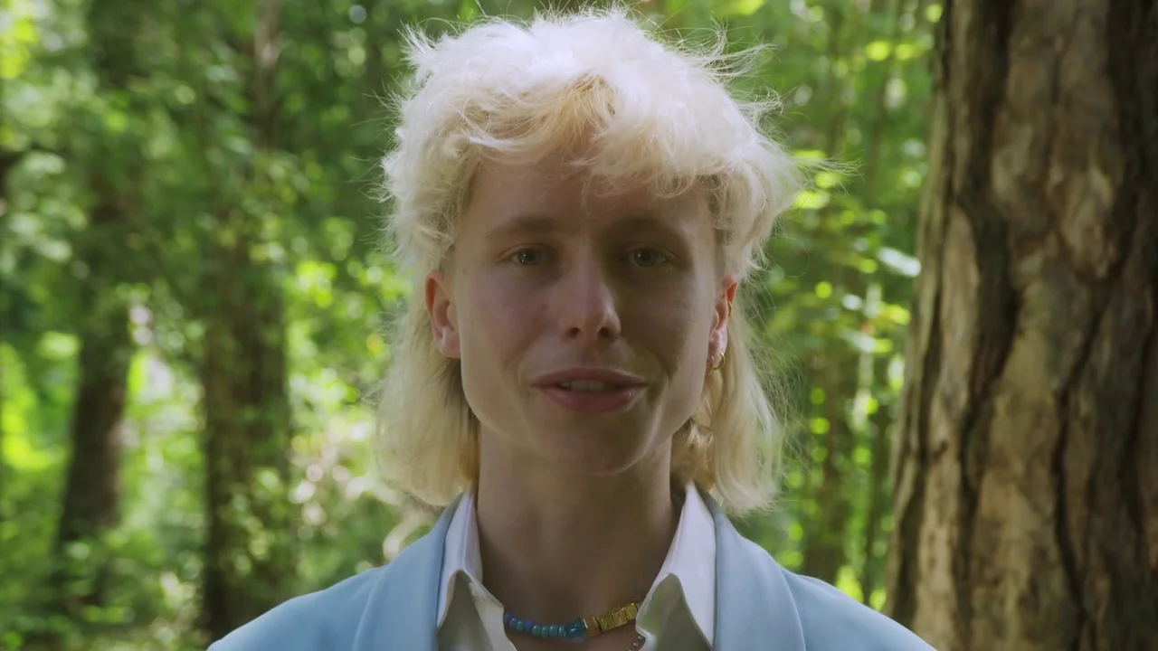 Orlando: My Political Biography (2023) movie still: closeup — A person stands in a sun-dappled forest setting with dense green trees in back. The…