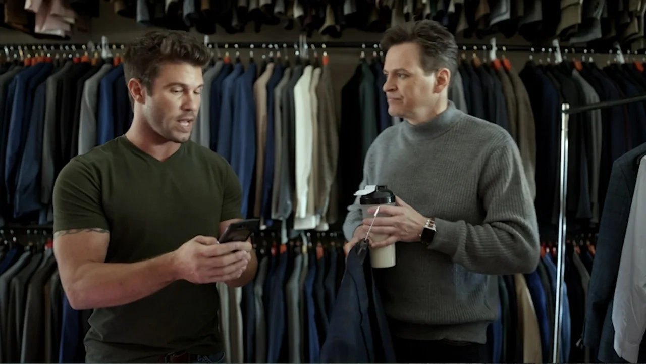 Ask Me to Dance (2022) movie still: medium — A well-organized men's clothing store with rows of hanging suits on both sides. Two men stand in the…
