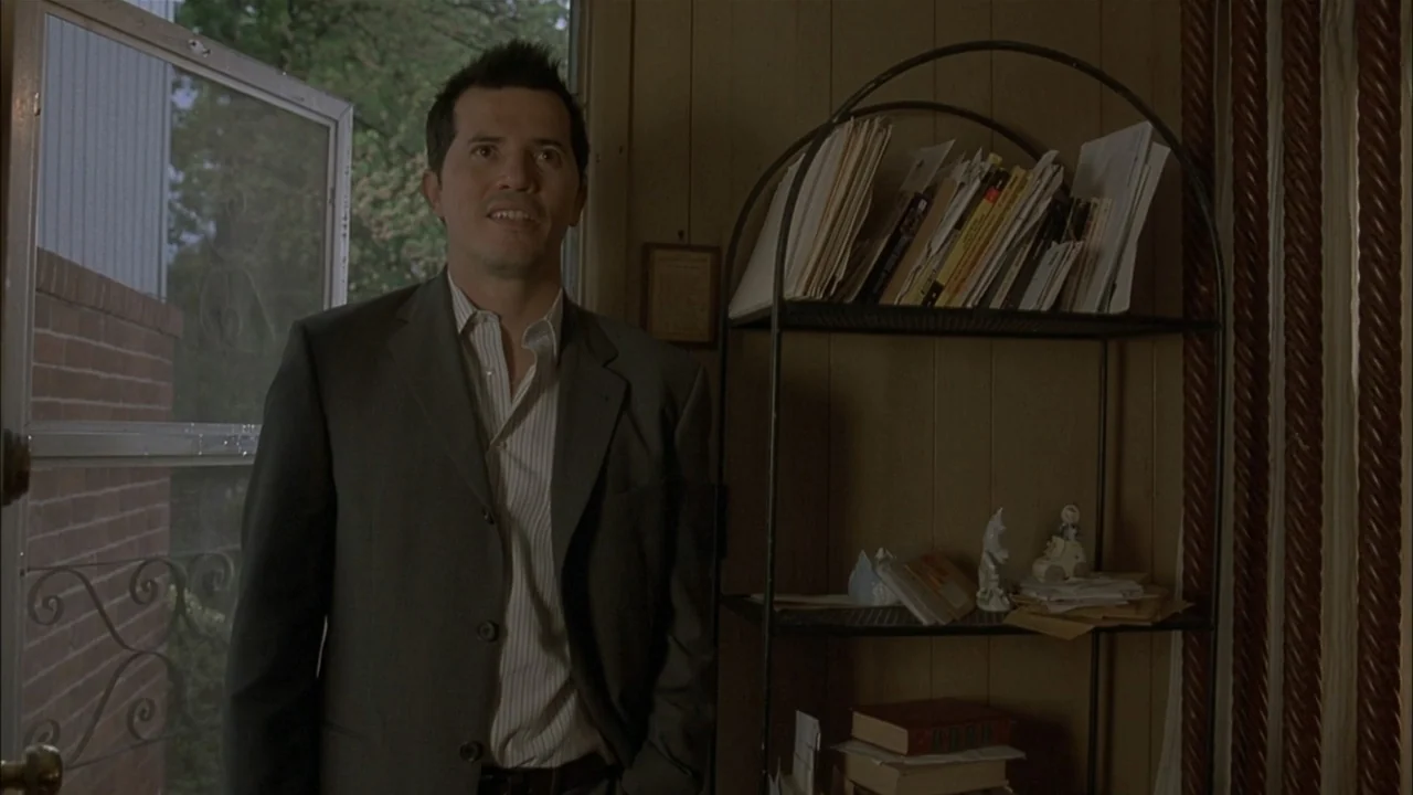 The Babysitters (2008) movie still: medium — A living room interior with wood-paneled walls and a metal wire shelving unit nearby. A man in a suit…