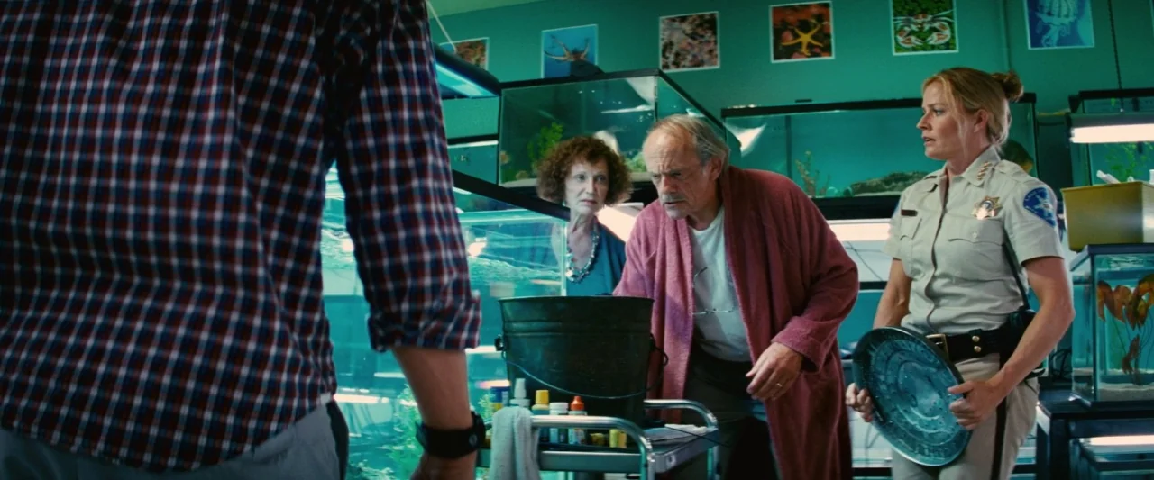 Piranha 3D (2010) movie still: medium — A pet shop with green walls features multiple fish tanks and framed posters. An elderly man in a robe…