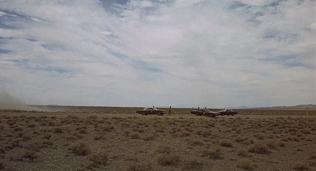 Vanishing Point (1971) movie still: wide — A vast, dry desert expanse under an expansive, cloudy sky with low brush. Several dark police cruisers…