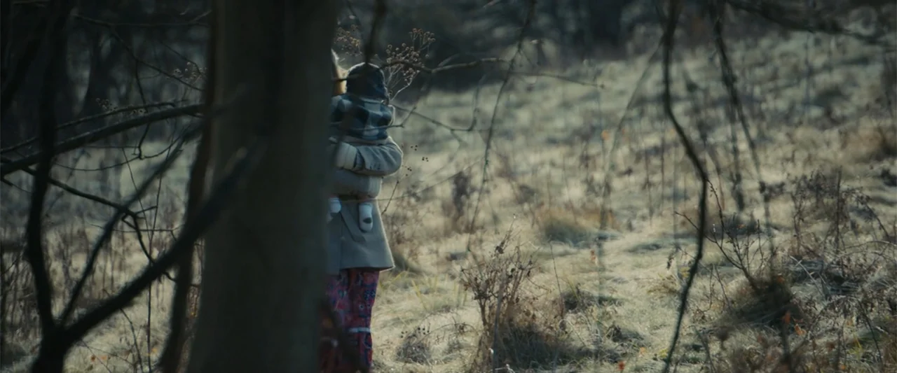 I'm Your Woman (2020) movie still: medium — A field of dried grasses and bare trees sits under overcast daylight conditions. A person carrying a…