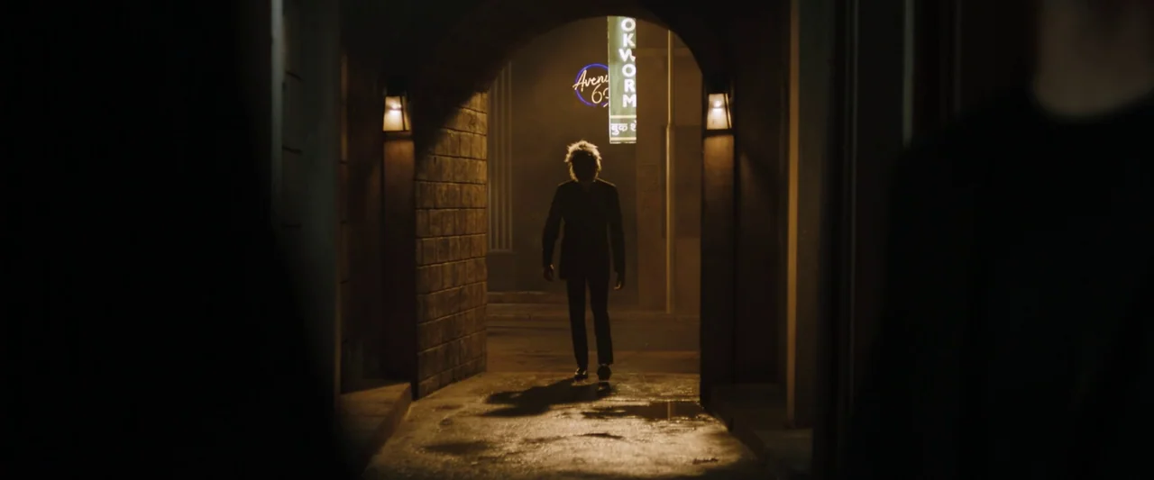Monkey Man (2024) movie still: fullbody — A dimly lit stone arched tunnel leads out to a nighttime street with neon signage. A person in a suit…