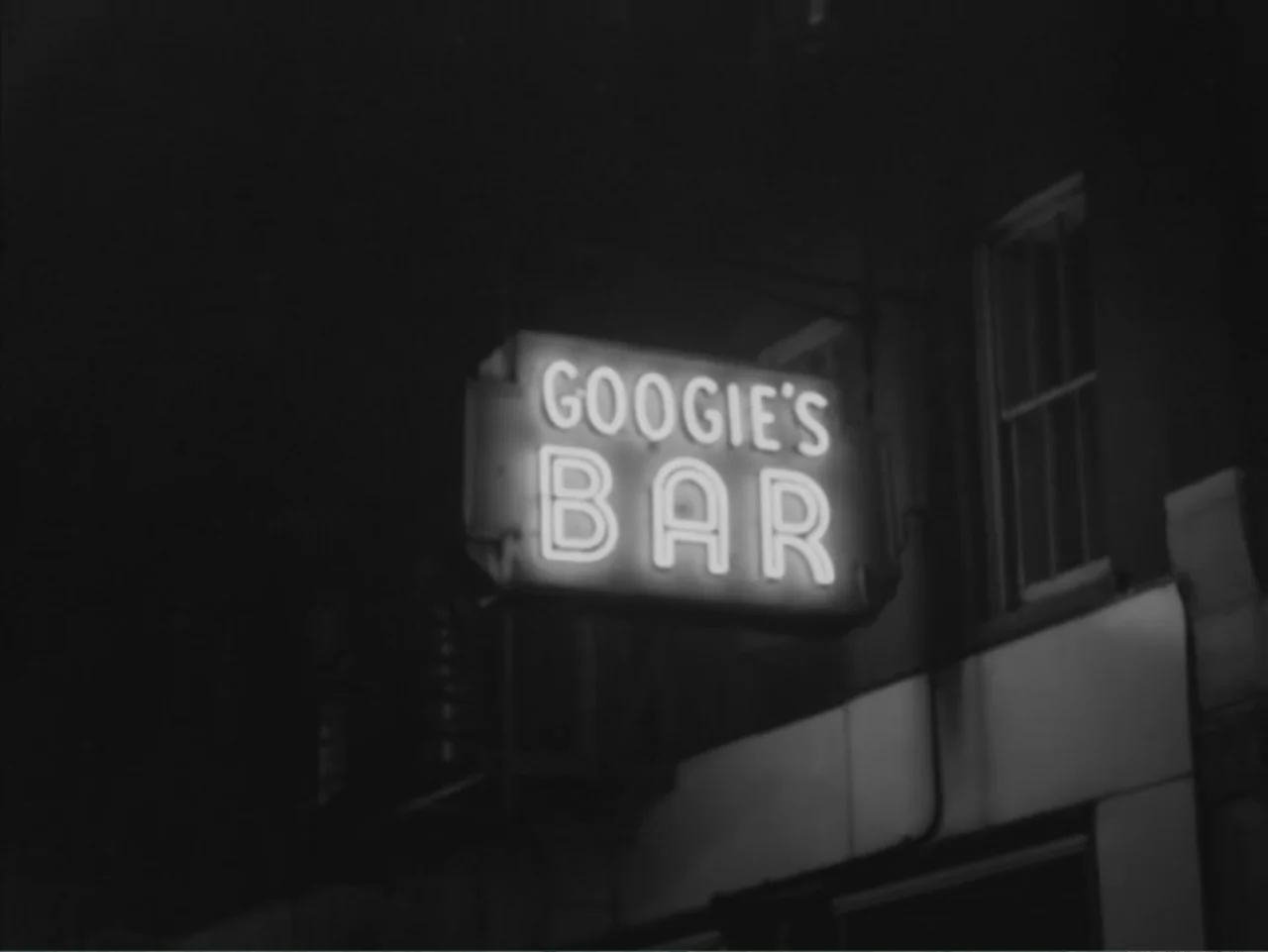Chained Girls (1965) movie still: wide — A dark city exterior building at night features a prominent glowing neon sign. The sign explicitly displays…