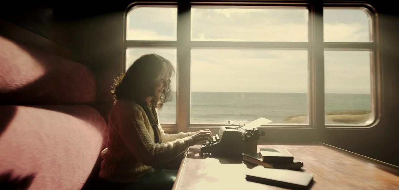 Peter Pan & Wendy (2023) movie still: medium — The interior of a train carriage features a window overlooking the ocean. A woman sits at a wooden…
