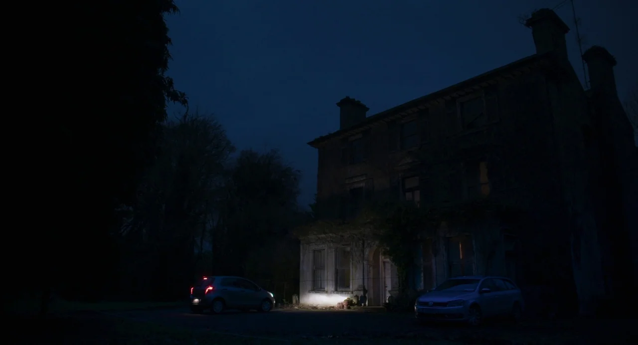 All You Need Is Death (2023) movie still: wide — A dark stone manor house sits under a clear night sky, obscured by shadows. Two parked cars occupy…