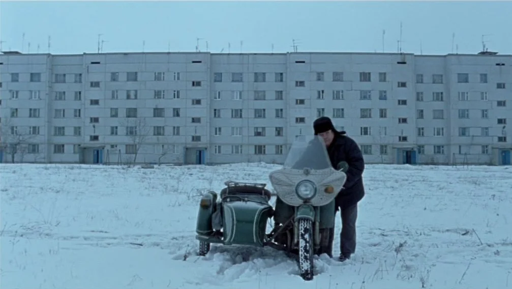 Import Export (2007) movie still: wide — A flat, snow-covered open space lies in front of a long, grey multi-story apartment block. A man in winter…