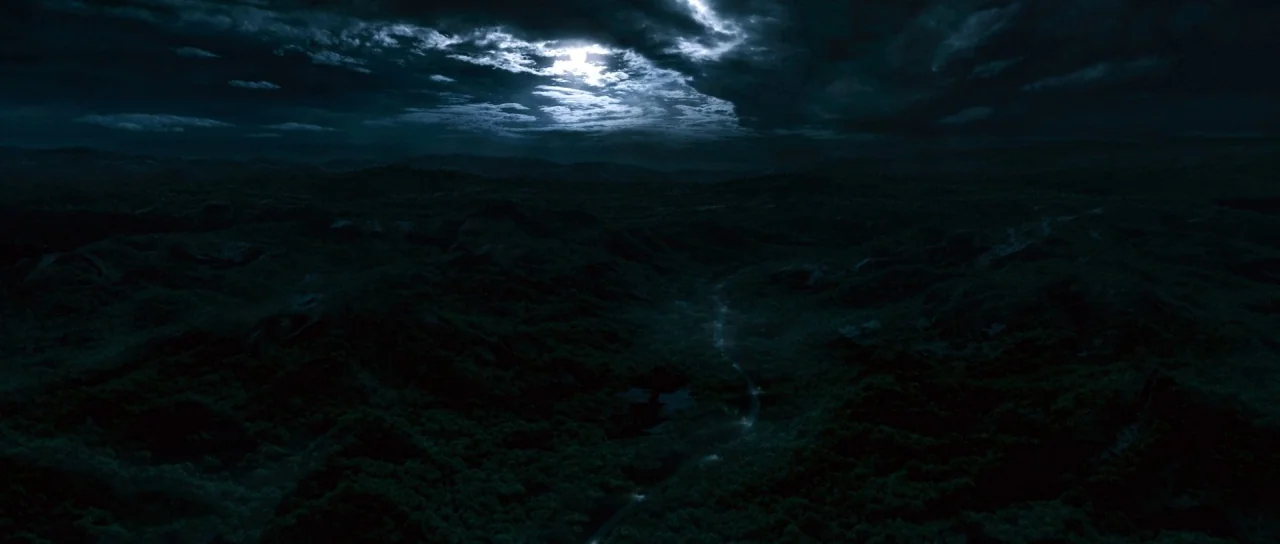 Silent Hill movie still: establishing — A vast landscape of dark, rolling hills under a cloudy night sky. A faint, reflective stream cuts through…