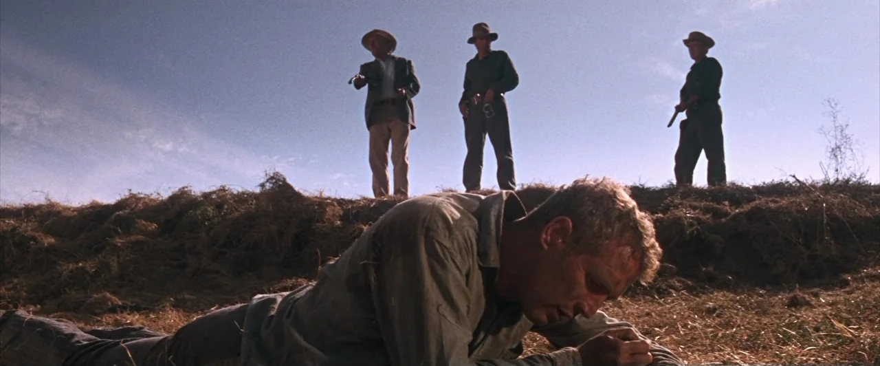 Cool Hand Luke (1967) movie still: wide — A dry, grassy field under a vast, clear blue sky during the day. A man lies on the ground while three men…