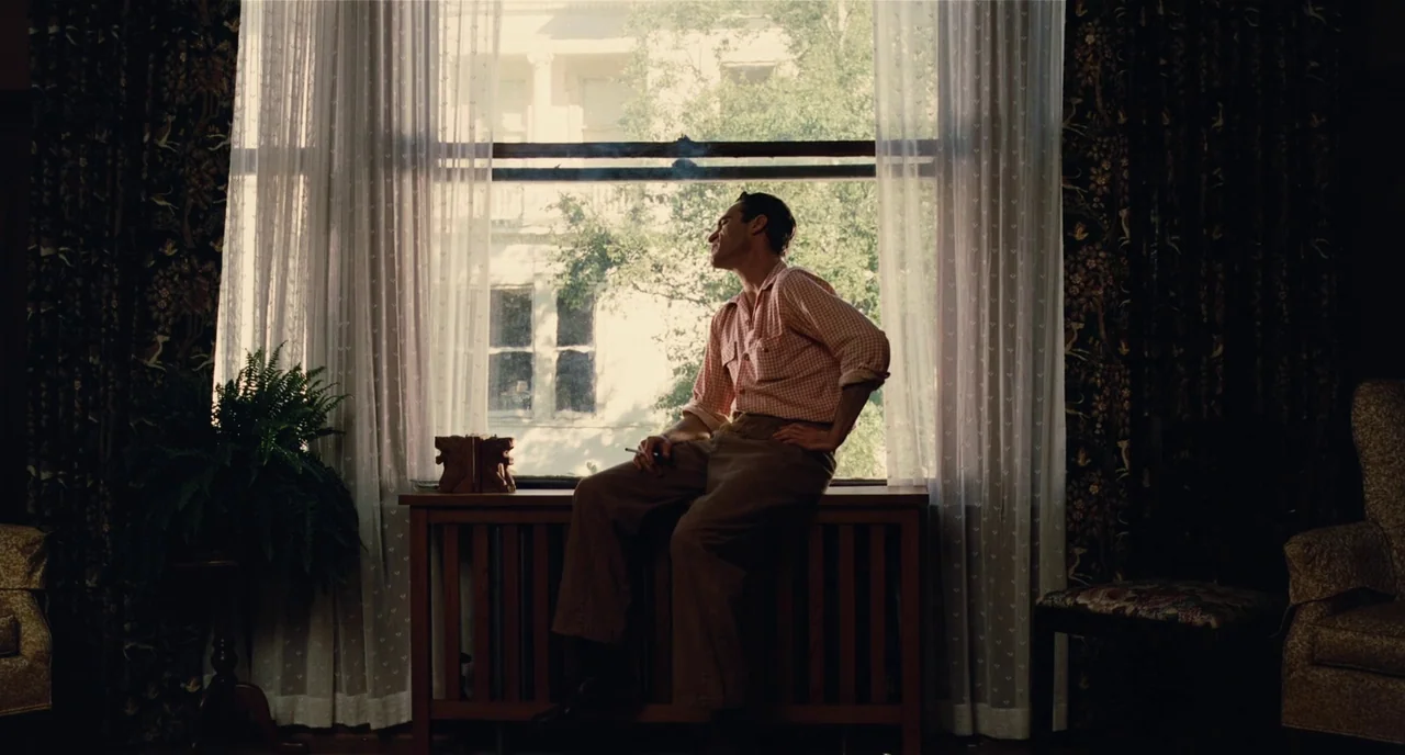 The Master (2012) movie still: fullbody — A 1940s living room features dark floral curtains and a radiator under a window. A man in a light-colored…