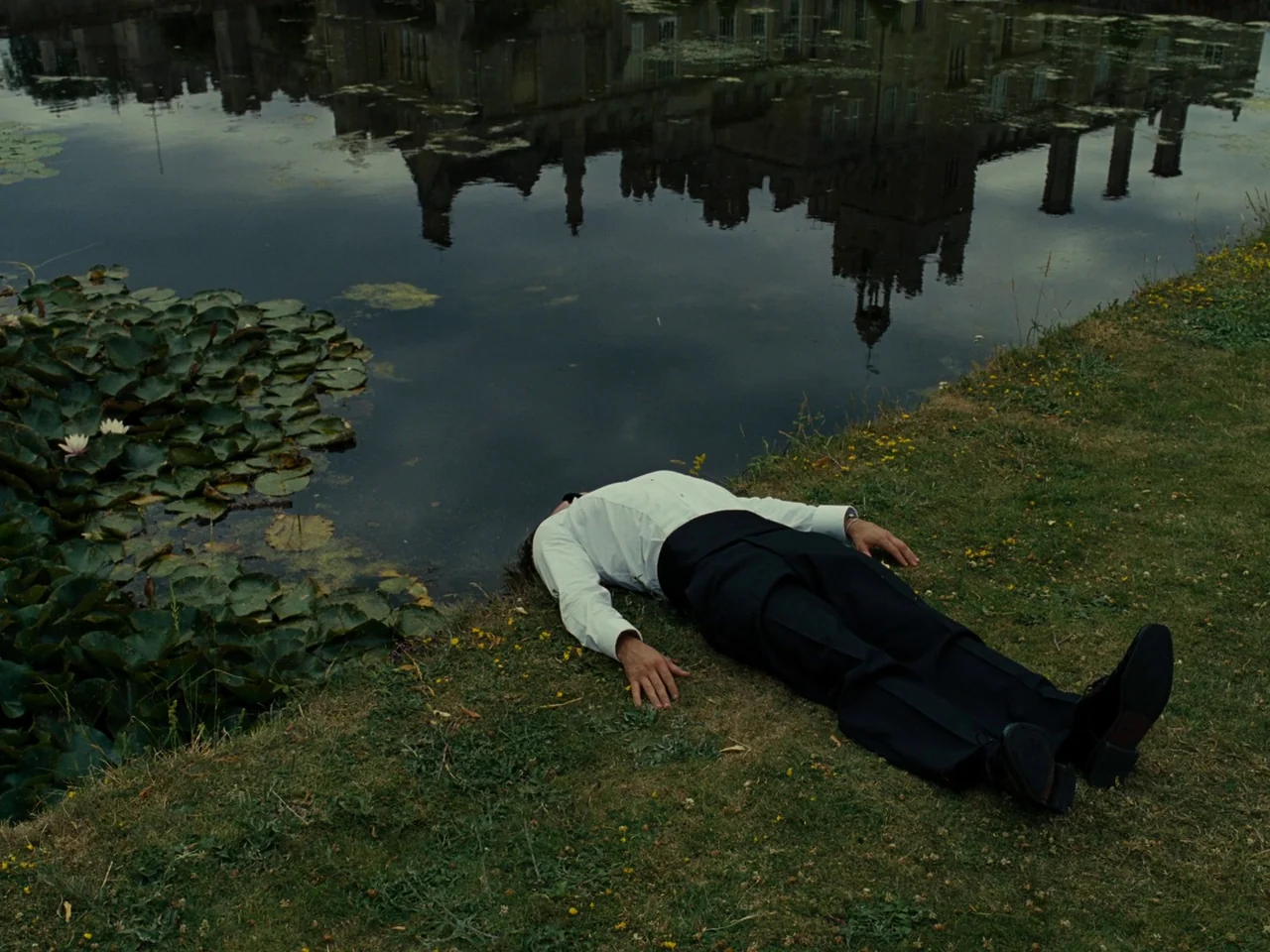 Saltburn (2023) movie still: fullbody — A man in formal attire lies on the grassy bank beside a dark pond. The water reflects a large stone building…