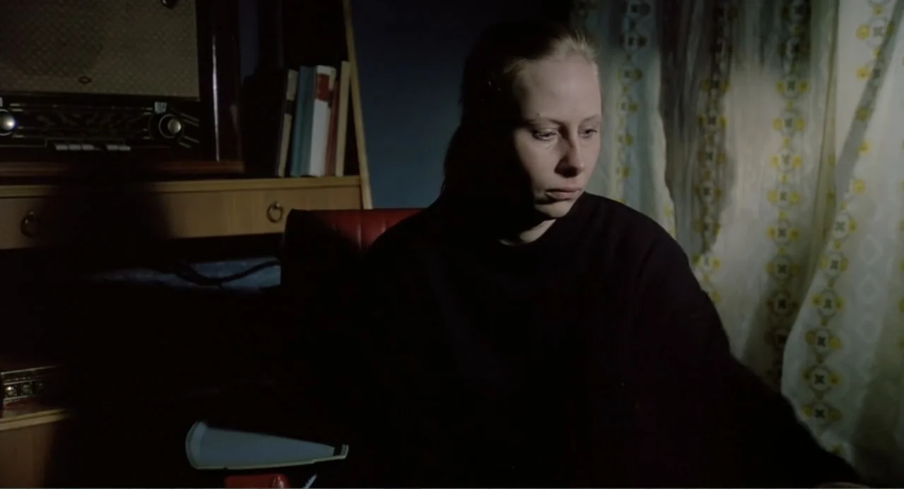 The Match Factory Girl (1990) movie still: medium — A dark indoor setting features a shelf with a vintage radio and books. A woman sits in a red…