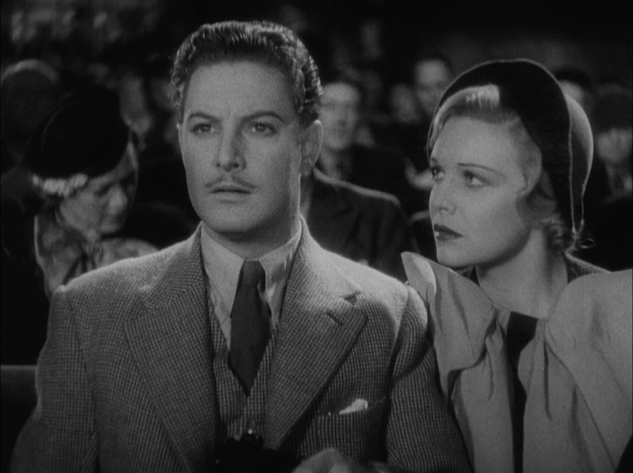 The 39 Steps (1935) movie still: medium — A dark, crowded public space with rows of seats occupied by an audience. A man and a woman sit closely…