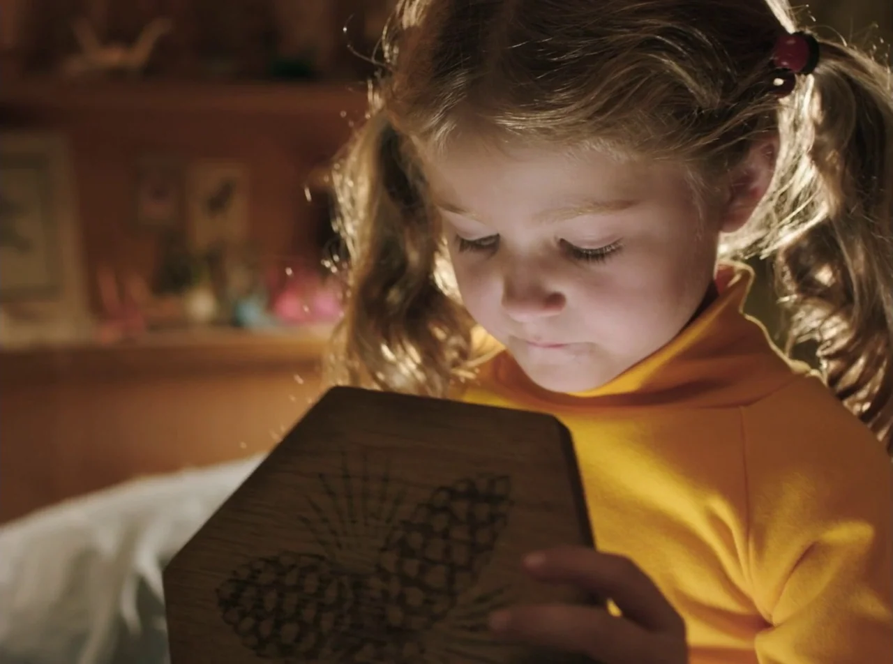 Girl Asleep (2015) movie still: closeup — A young girl in a yellow shirt looks at an illuminated wooden object. The background is a dark,…
