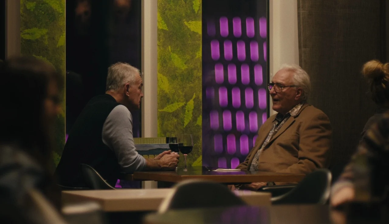 Land Ho! (2014) movie still: medium — A dimly lit room features two men sitting at a table with wine glasses. One man in a tan blazer faces the…