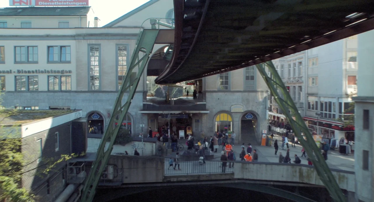 Pina (2011) movie still: wide — A Wuppertal Suspension Railway track arches over a busy pedestrian station and city buildings. People move through…