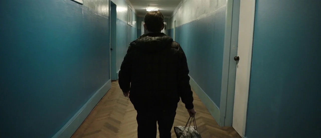 The Tribe (2014) movie still: medium — A long corridor features blue painted walls and a light brown chevron pattern floor. A man walks away from…